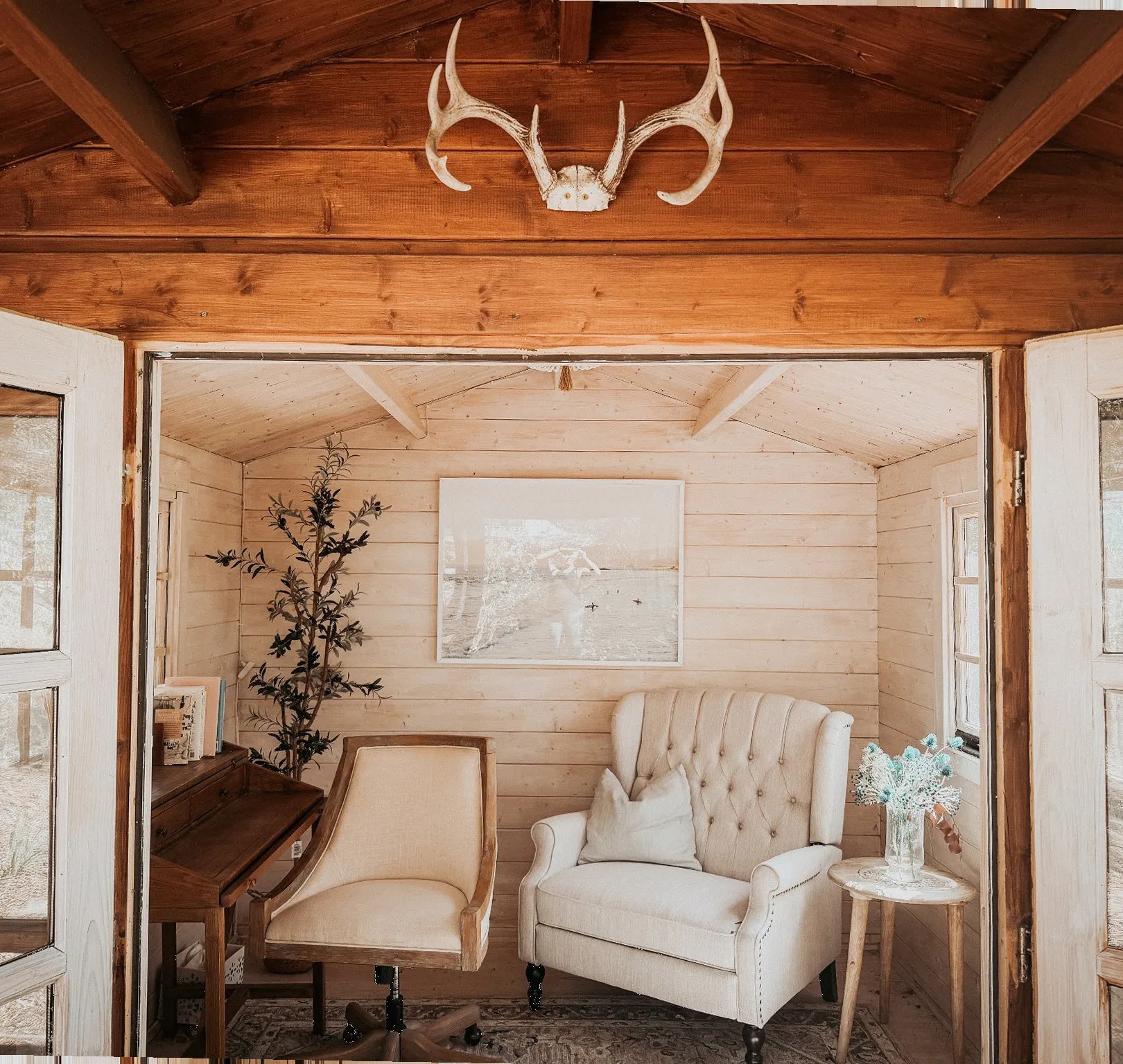 Inside a small wooden writing shack: a writing desk and chair, a tufted armchair, an olive tree and antlers above open glass doors